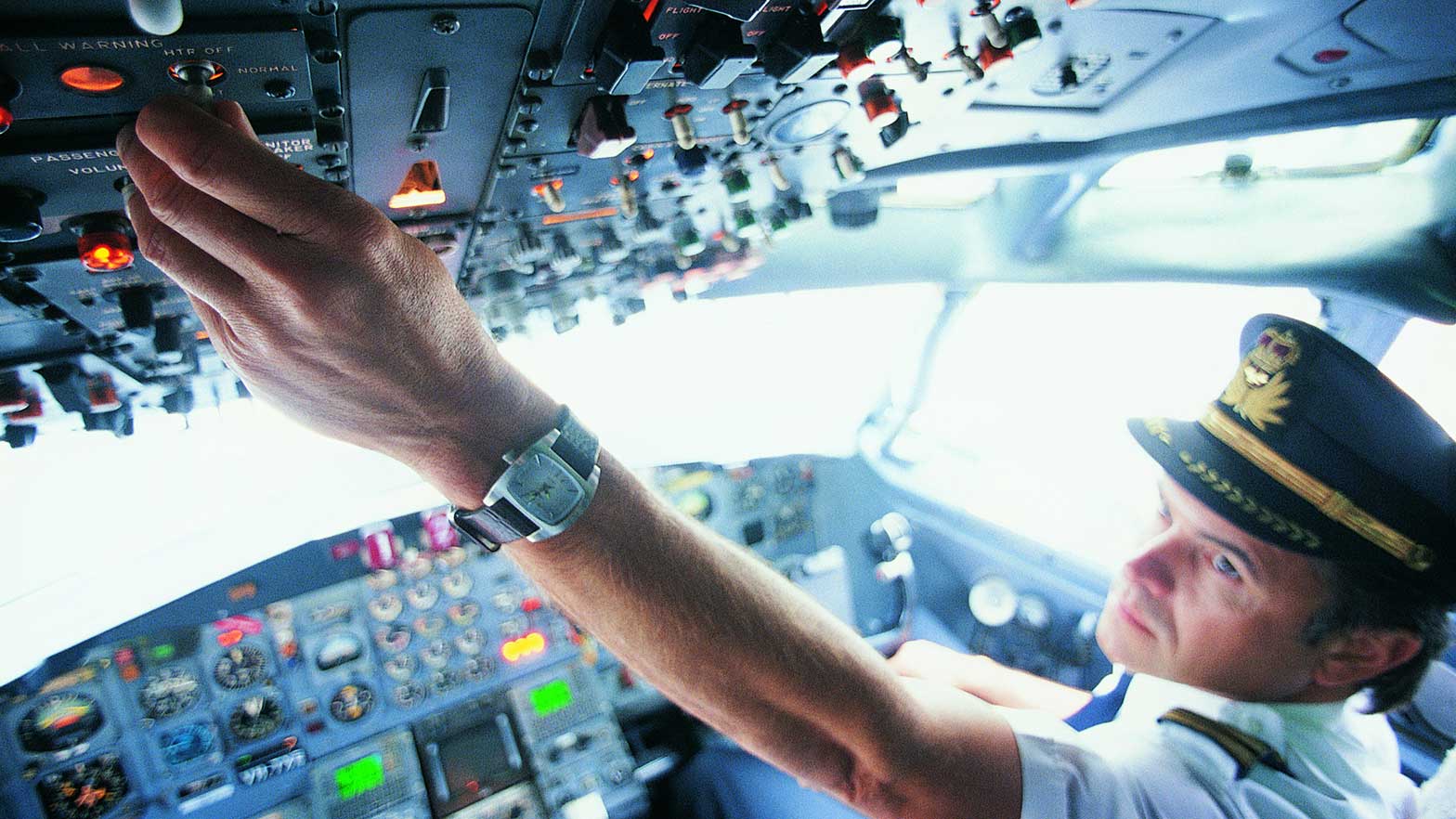 A commercial airline pilot in the cockpit flips a switch on a panel of instruments above him 
