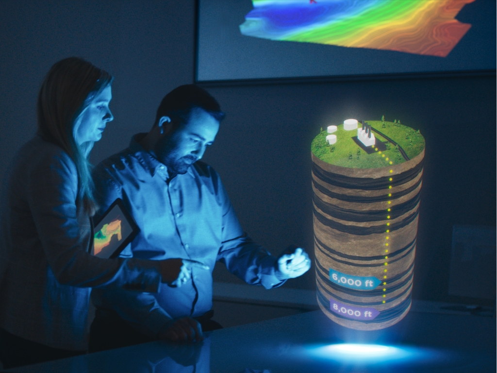 In a dark office room, two engineers study a projection mapping of a carbon capture site that shows the Earth’s layers and drilling depth.