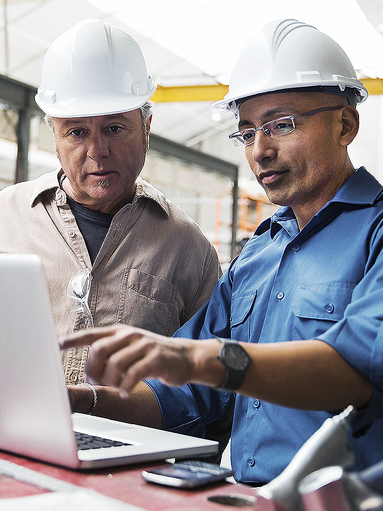 Two workers in a manufacturing plant looking at an open laptop. They both wear hard hats and there are tools on the table.