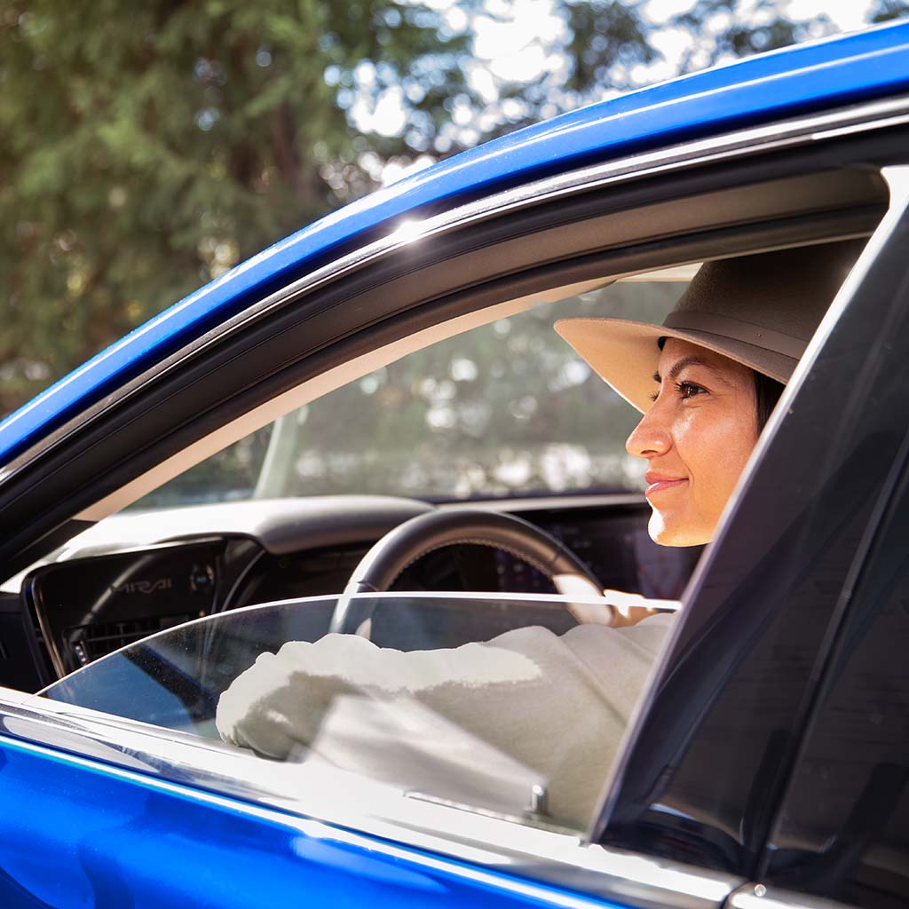 A young woman wearing a wide brimmed hat looks cheerfully through her partially open window from the driver's seat of a blue sedan