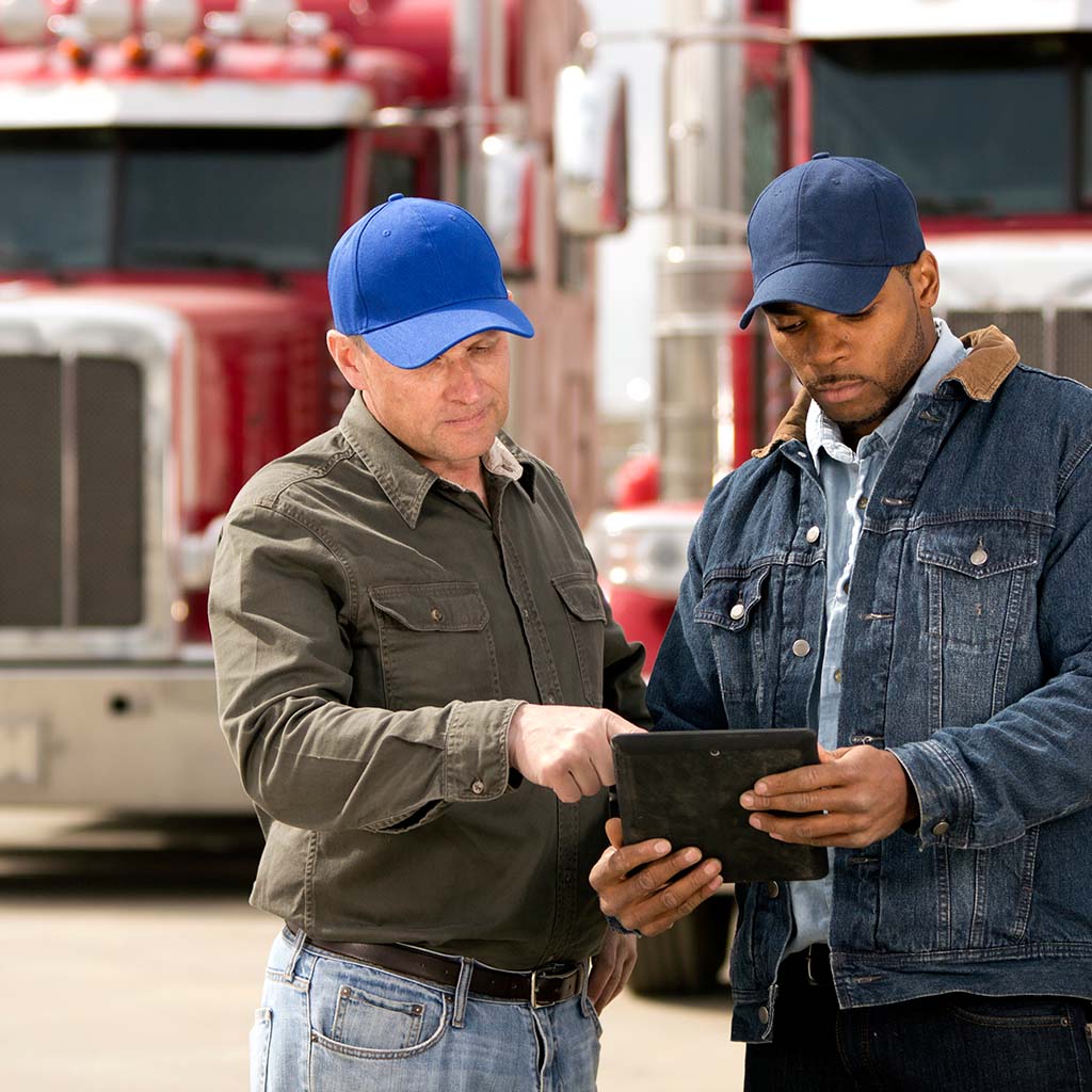Two denim-wearing truck drivers check information on their tablet, their red big rigs parked in the background
