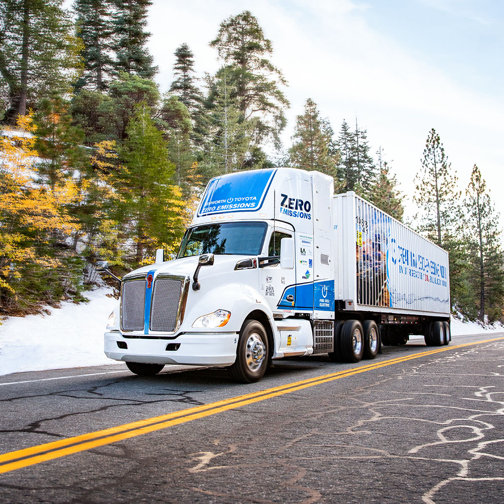 A white modern hydrogen eighteen wheeler with blue insignia drives on a two lane in the PNW. Snow covers the hillsides
